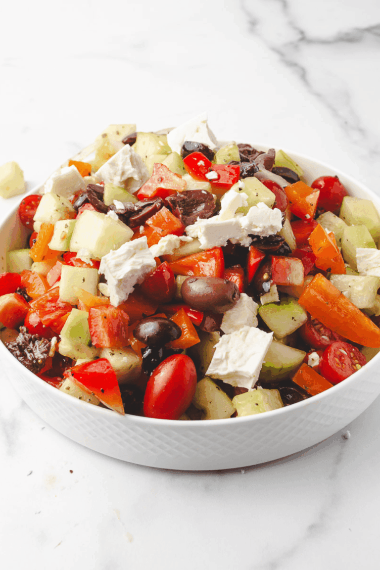 Classic Greek salad recipe served in a white diamond-textured bowl on a white marble surface, topped with large chunks of hand-crumbled feta cheese, Kalamata olives, grape tomatoes, diced cucumbers and colorful red and orange bell peppers.