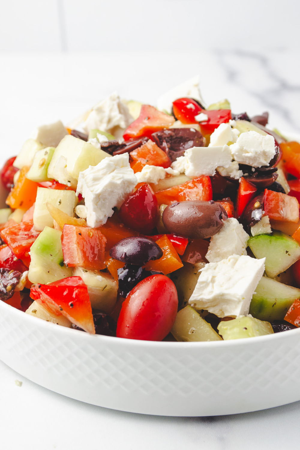 Greek salad recipe in a white diamond-textured bowl on a white marble surface, filled with diced cucumbers, grape tomatoes, chopped red and orange bell peppers, Kalamata olives and large chunks of hand-crumbled feta cheese.