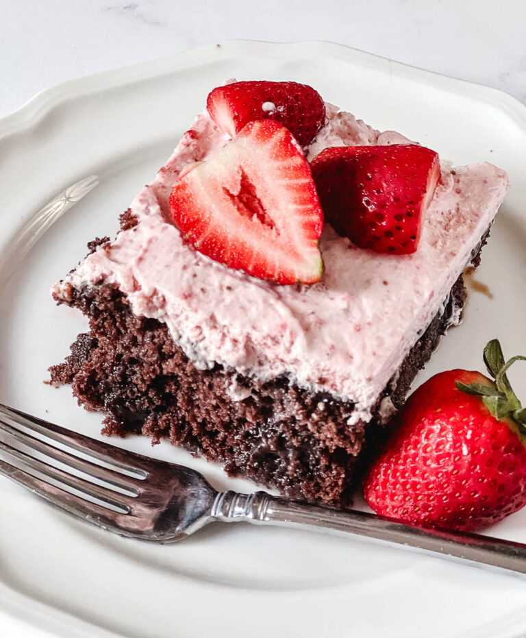 A slice of chocolate strawberry poke cake with pink strawberry whipped cream frosting and fresh halved strawberries on top, served on a white plate with a fork and a whole strawberry on the side.