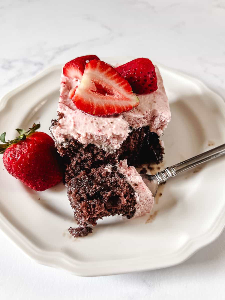 A slice of chocolate strawberry poke cake on a white plate with a fork taking the first bite, topped with pink strawberry whipped cream frosting and fresh halved strawberries, with a whole strawberry on the side.