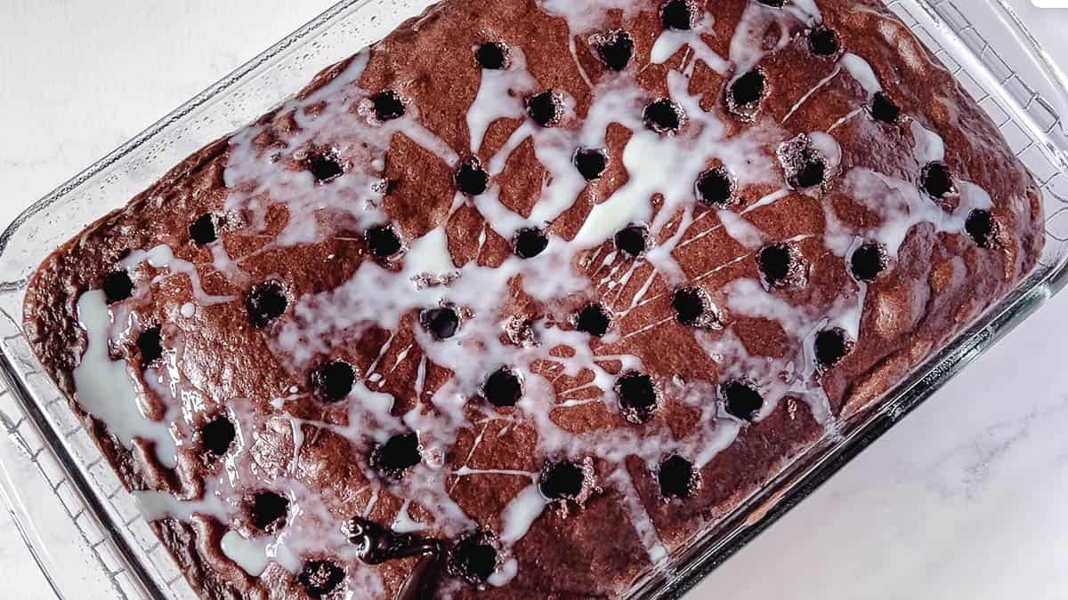 Overhead view of a chocolate poke cake in a glass 9x13 pan with holes poked across the surface and sweetened condensed milk poured over the top, soaking into every hole.