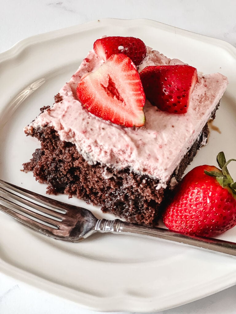 A slice of chocolate strawberry poke cake with pink strawberry whipped cream frosting and fresh halved strawberries on top, served on a white plate with a fork and a whole strawberry on the side.