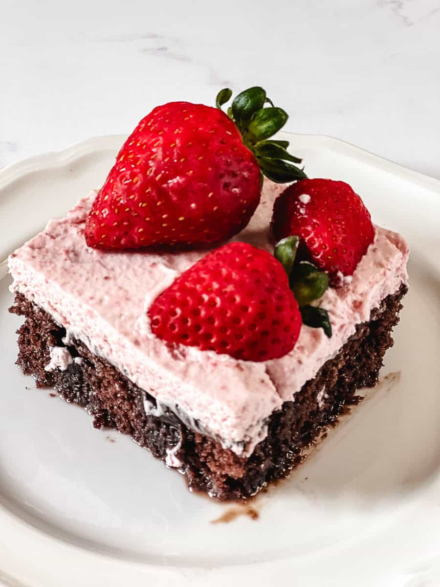 A slice of chocolate strawberry poke cake on a white plate topped with pink strawberry whipped cream frosting and three whole fresh strawberries with stems on, on a white marble surface.