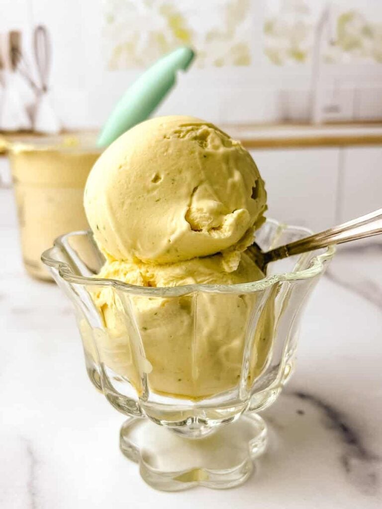 Ninja Creami dairy free ice cream scooped into a clear glass dessert dish on a white marble surface, with a second serving of mango coconut lime ice cream and a teal spoon visible in the background.