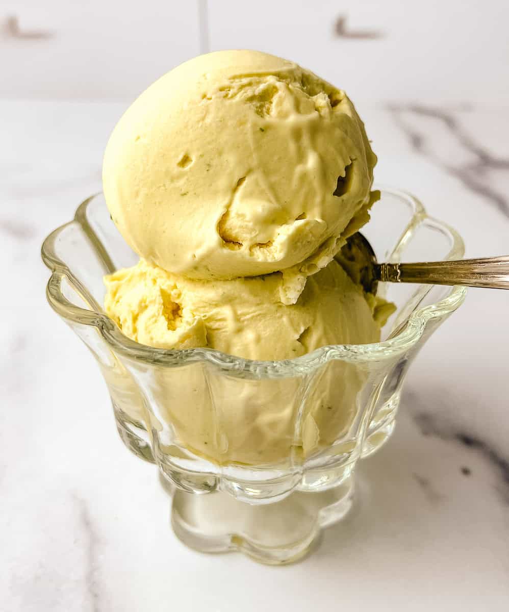 Two scoops of Ninja Creami mango coconut lime ice cream in a clear glass dessert dish with a silver spoon on a white marble surface, showing the creamy dairy free texture.