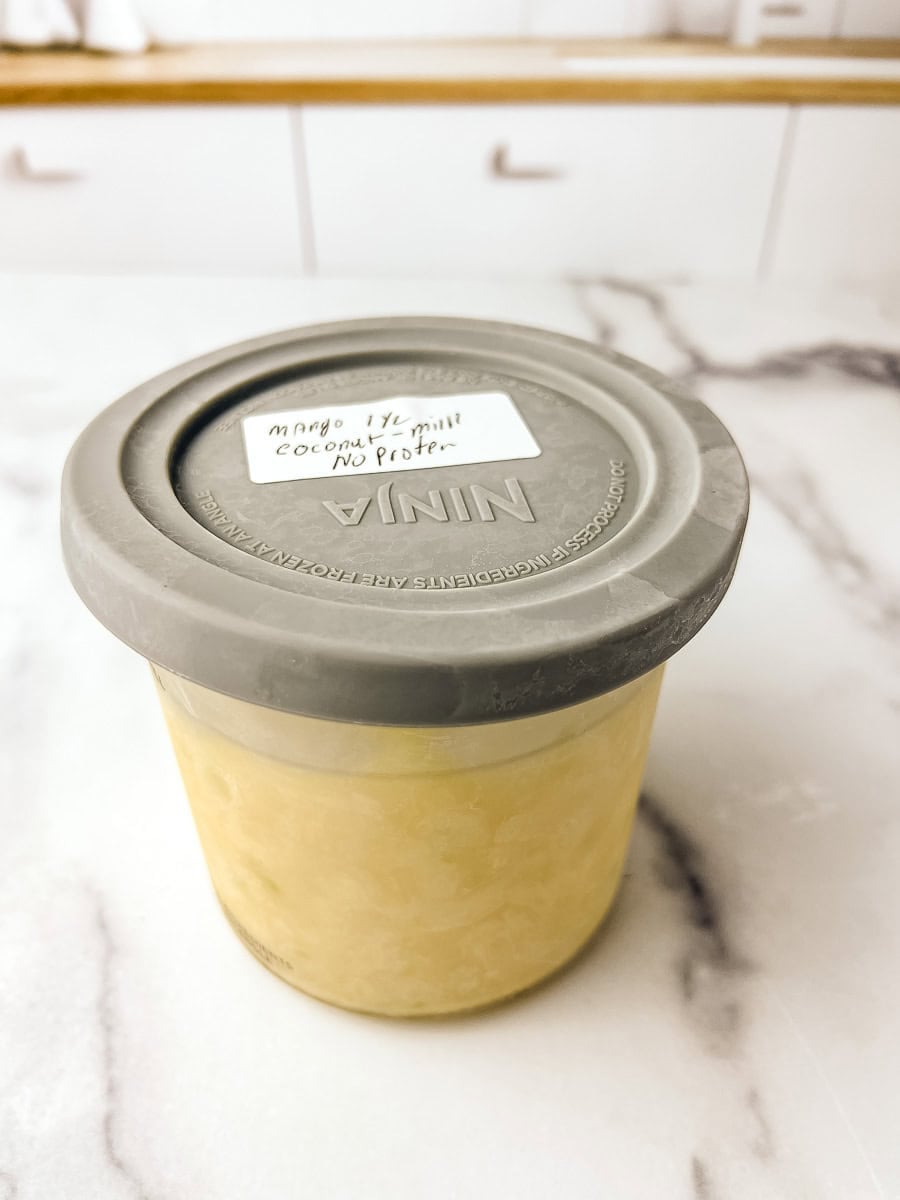 Ninja Creami pint container filled with frozen mango coconut lime dairy free ice cream mixture, labeled and ready to spin, sitting on a white marble surface.