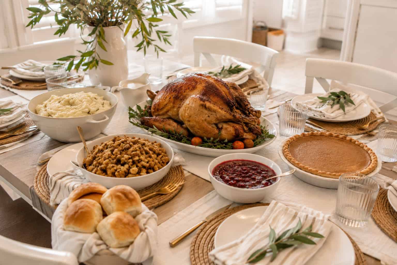 Modern Thanksgiving dinner table with a roasted turkey, mashed potatoes, stuffing, cranberry sauce, rolls, and pumpkin pie on a light wood table in a bright, airy farmhouse kitchen with soft natural light.