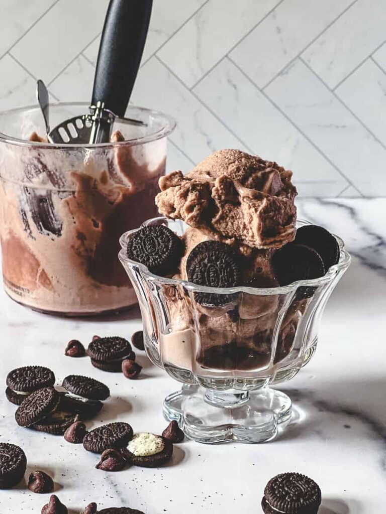 Scoops of creamy chocolate Ninja Creami ice cream topped with Oreos in a glass bowl, with more Oreos and chocolate chips scattered on the white marble surface, and the Creami machine blurred in the background.