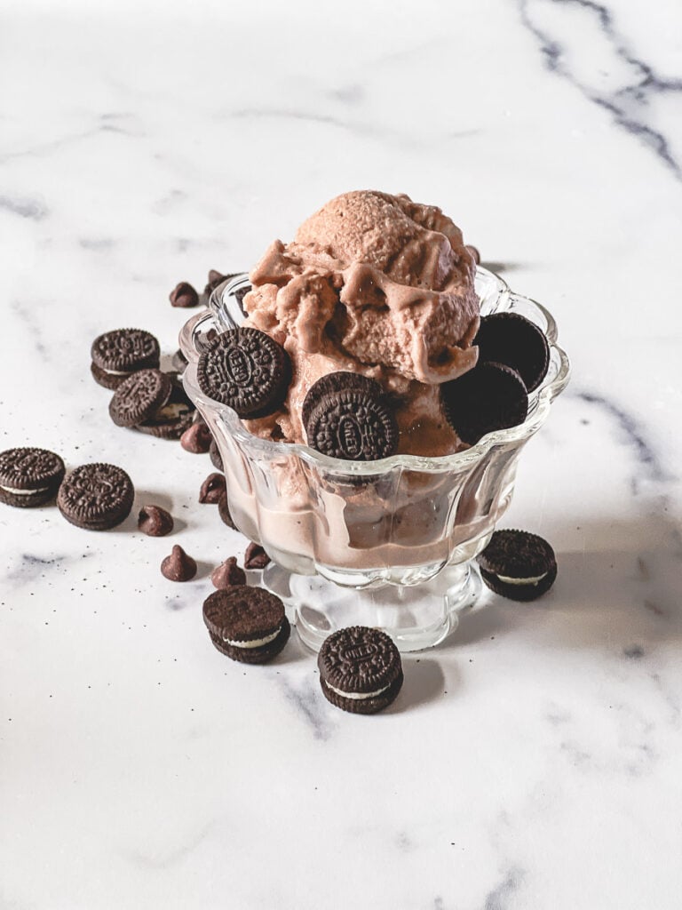 Creamy chocolate Ninja Creami ice cream piled high in a pedestal glass bowl, topped with Oreo cookies and chocolate chips, with more Oreos and chocolate chips scattered on the marble surface.