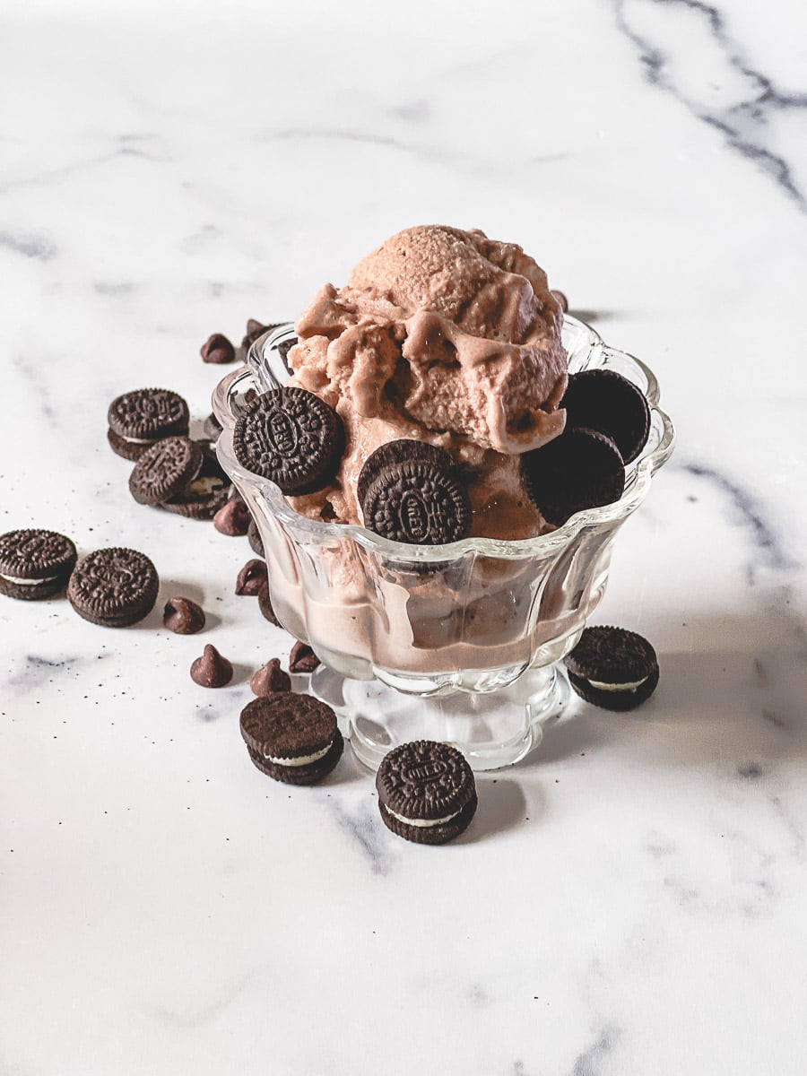 Creamy chocolate Ninja Creami ice cream piled high in a pedestal glass bowl, topped with Oreo cookies and chocolate chips, with more Oreos and chocolate chips scattered on the marble surface.