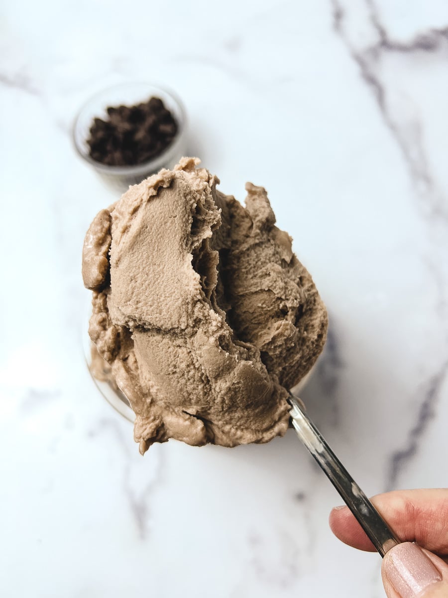 A spoon holding two generous scoops of creamy chocolate Ninja Creami ice cream, with a small bowl of chocolate chips blurred in the background on a marble surface.