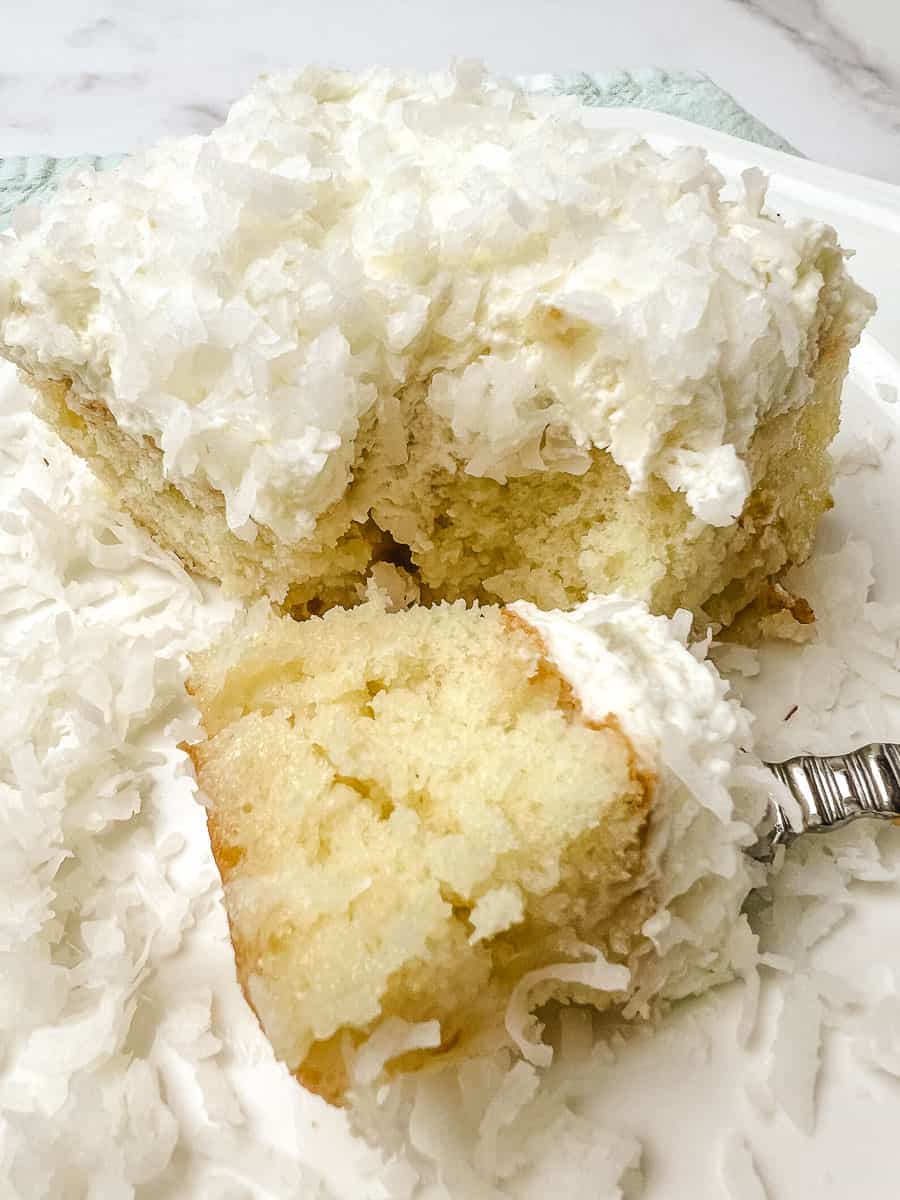 Forkful of coconut poke cake resting beside the slice on a white plate, showing the moist crumb under thick whipped cream and shredded coconut.
