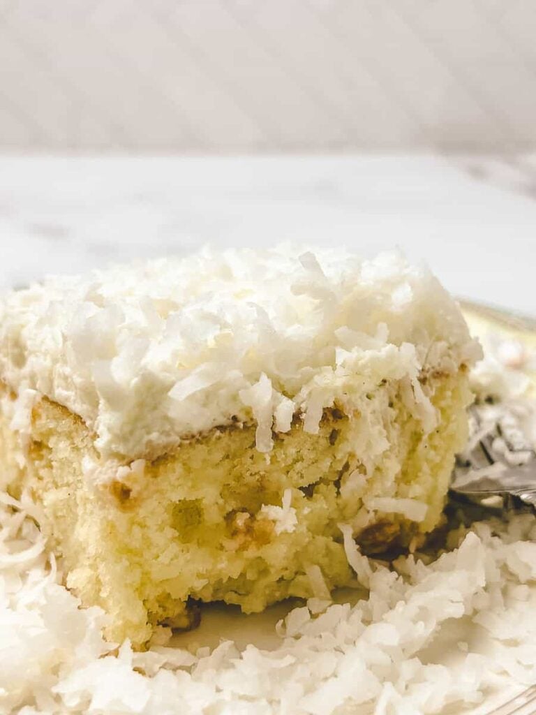 Side view of a coconut poke cake slice showing the soaked streaks running through the crumb, topped with whipped cream and shredded coconut.