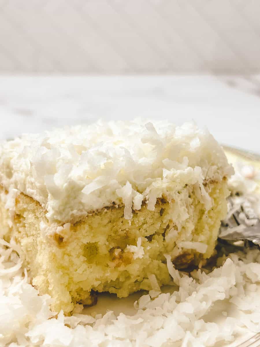 Side view of a coconut poke cake slice showing the soaked streaks running through the crumb, topped with whipped cream and shredded coconut.