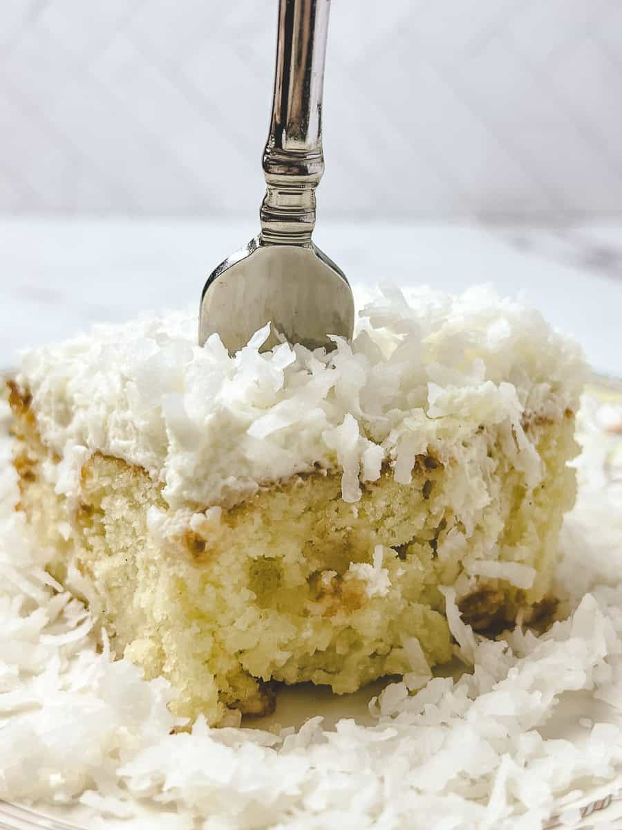 Square slice of coconut cream poke cake showing the moist soaked crumb inside, topped with whipped cream and shredded coconut on a clear glass plate with a vintage silver fork.