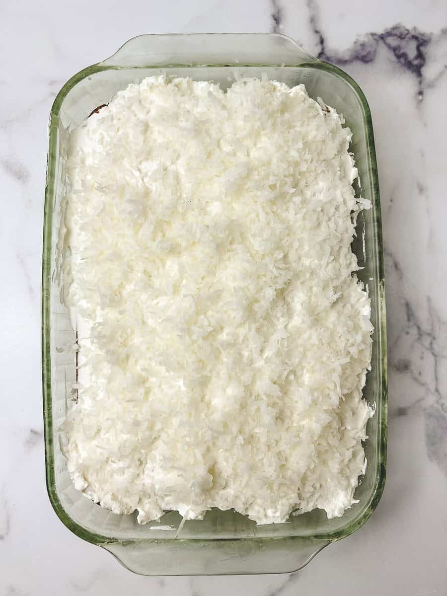 Whole coconut cream poke cake in a green glass baking dish, covered edge to edge with whipped cream and shredded coconut, on a white marble counter.