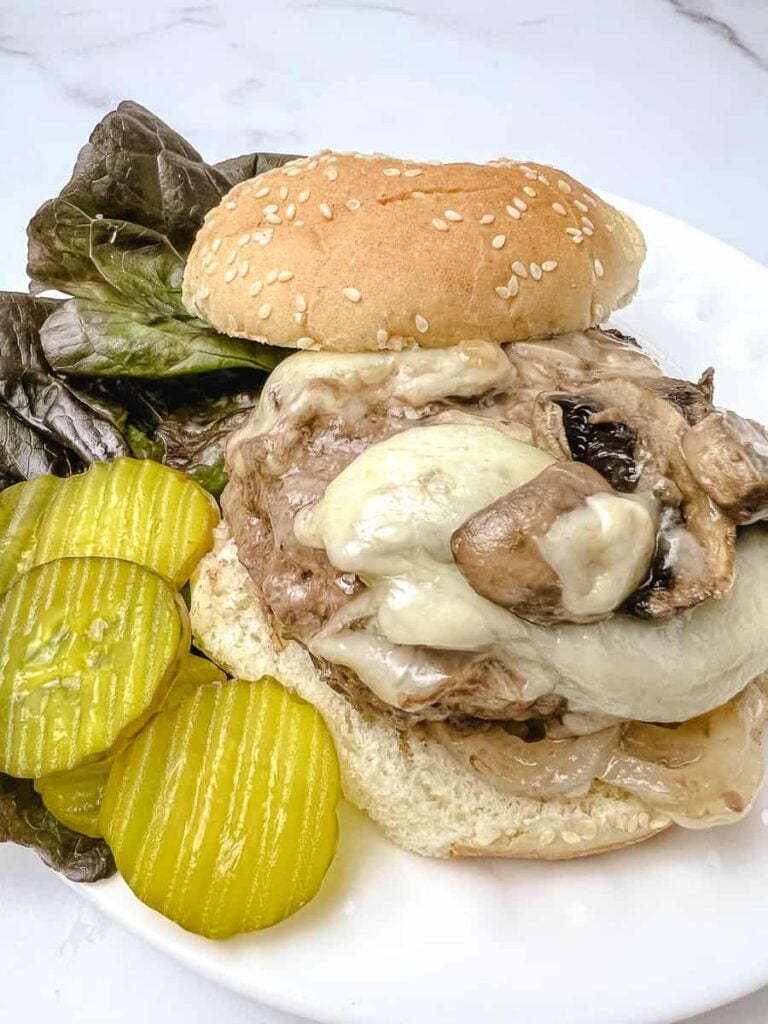 Close up of a baked mushroom swiss burger with melted swiss cheese and mushrooms on a sesame seed bun, served on a white plate with pickle chips and lettuce on a white marble counter