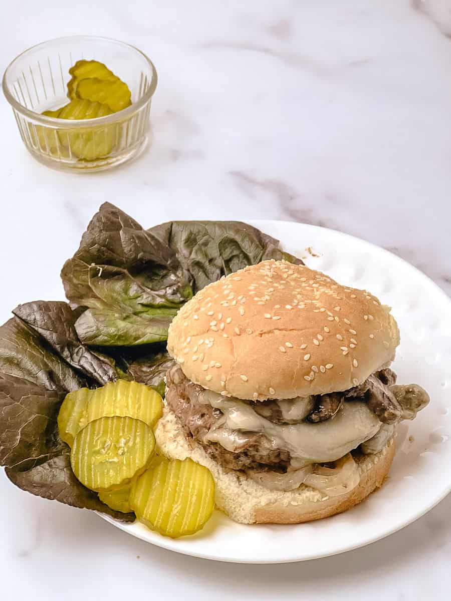 Baked mushroom swiss burger with melted swiss cheese on a sesame seed bun, served on a white plate with lettuce and pickle chips, small glass bowl of pickles in the background on a white marble counter