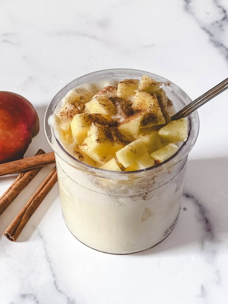 A Ninja Creami pint of caramel apple pie protein ice cream topped with fresh apple chunks and cinnamon, with a spoon, a red apple, and cinnamon sticks on a white marble counter.