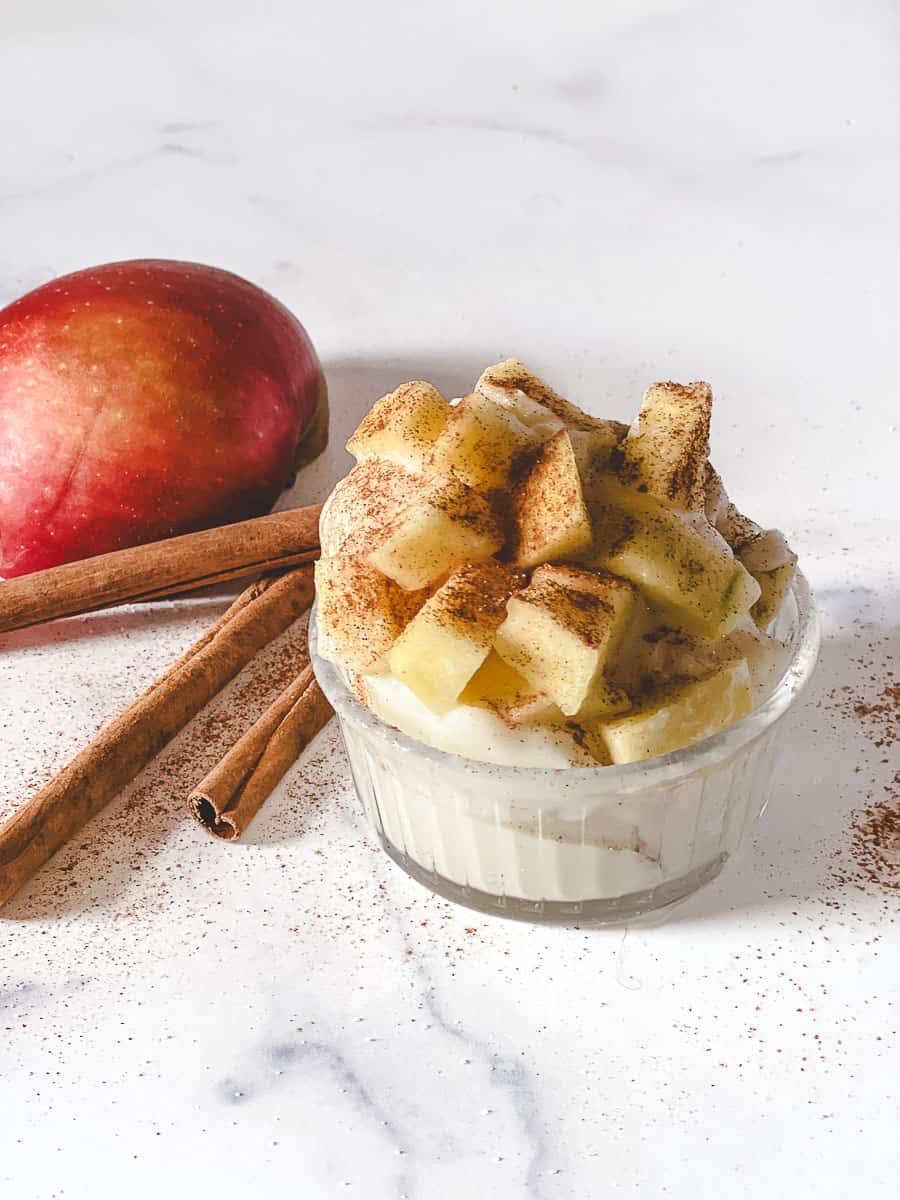 A glass dish of caramel apple pie protein ice cream topped with apple chunks and cinnamon, with a red apple and cinnamon sticks on marble.