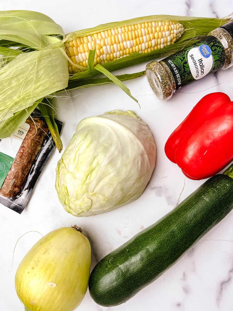 Raw ingredients for an easy cabbage recipe laid out on a white marble counter, including fresh corn on the cob, a whole head of cabbage, red bell pepper, zucchini, a yellow onion, ground beef, and a jar of Italian seasoning, everything you need for a low carb dinner