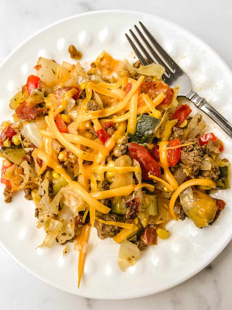 Easy cabbage recipe served on a white scalloped plate, piled with melted cheddar cheese, ground beef, red bell peppers, corn, and chopped zucchini, a fork resting beside it on a white marble counter, a colorful low carb dinner ready to eat