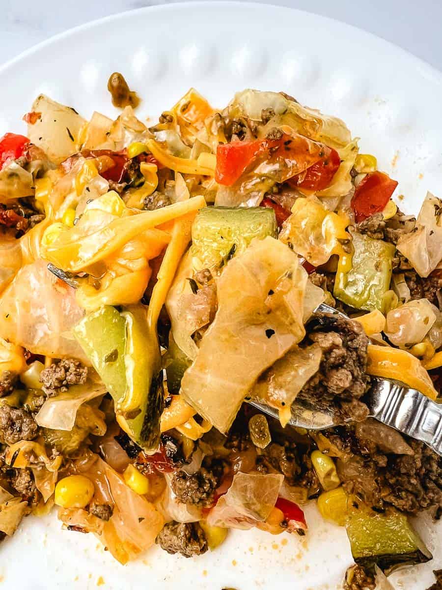 Close up of a fork lifting a bite of easy cabbage recipe from a white scalloped plate, showing tender cabbage, ground beef, zucchini, and melted cheddar, a hearty low carb dinner ready to eat