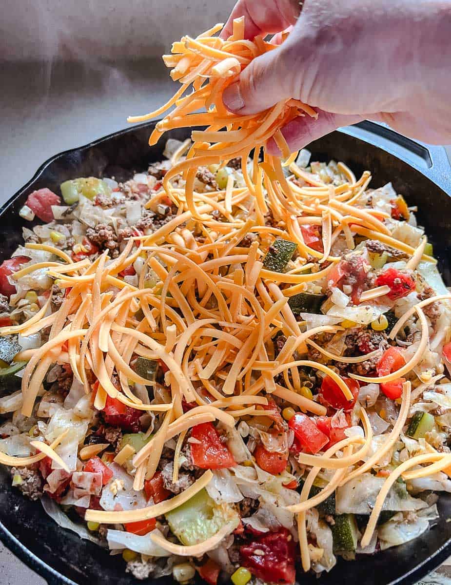 Shredded cheddar cheese being sprinkled by hand over an easy cabbage recipe cooking in a cast iron skillet, with ground beef, red peppers, corn, and zucchini visible, the final step in this one pan low carb dinner