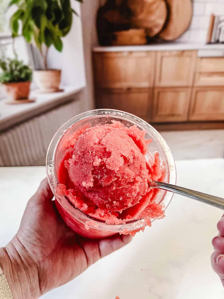 Hand holding a Ninja Creami pint of freshly spun strawberry sorbet with a spoon, in a kitchen with wood cabinets and potted plants on the windowsill.