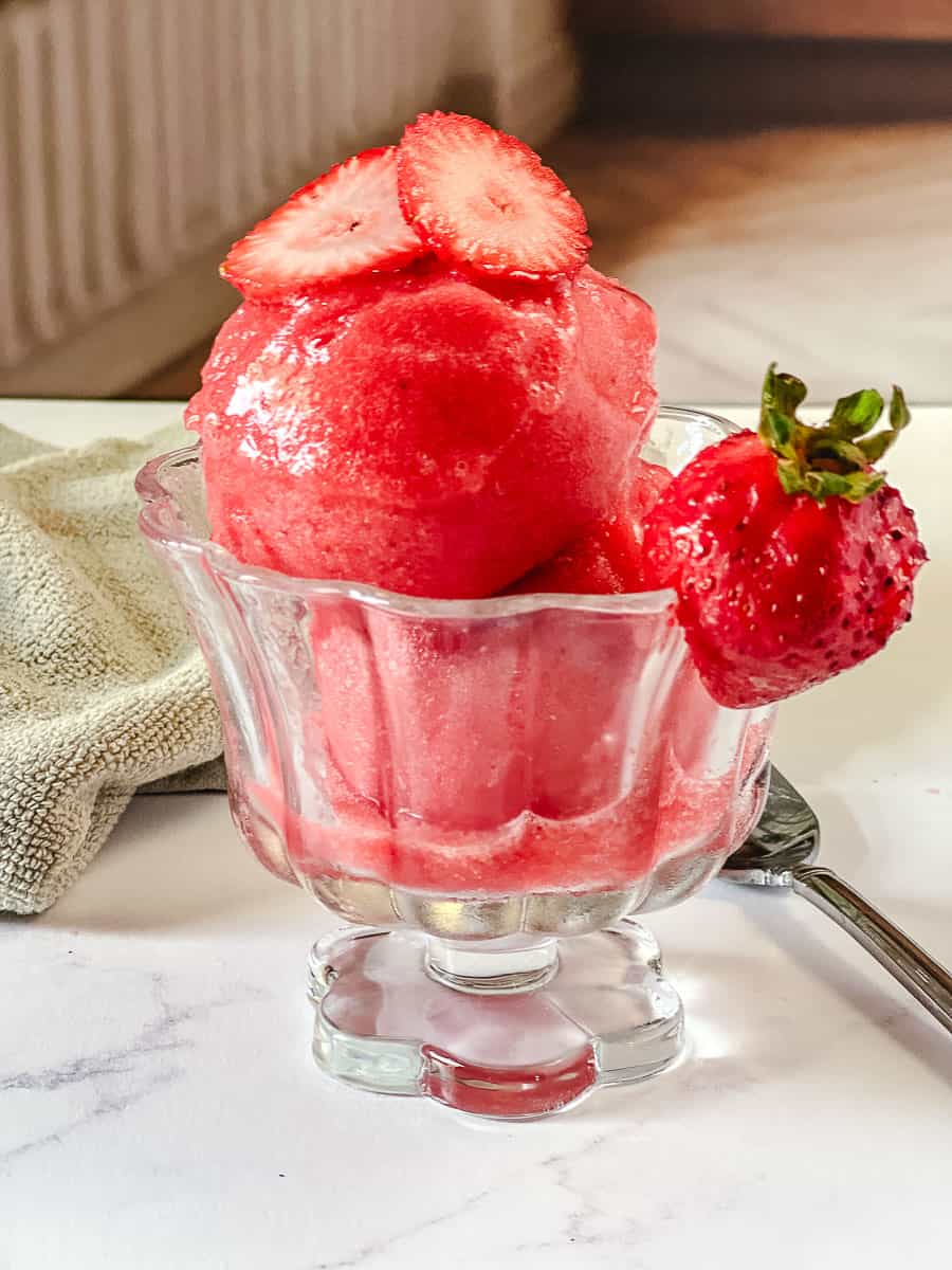 Glass footed dish of pink Ninja Creami strawberry sorbet, topped with sliced strawberries and a whole strawberry, on a white marble counter with a tan towel and spoon in the background.