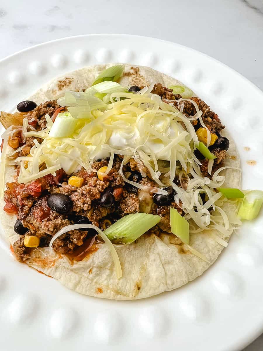 Taco skillet dinner with ground beef, black beans, and corn served on a soft flour tortilla topped with shredded cheese, sour cream, and green onions on a white ridged plate
