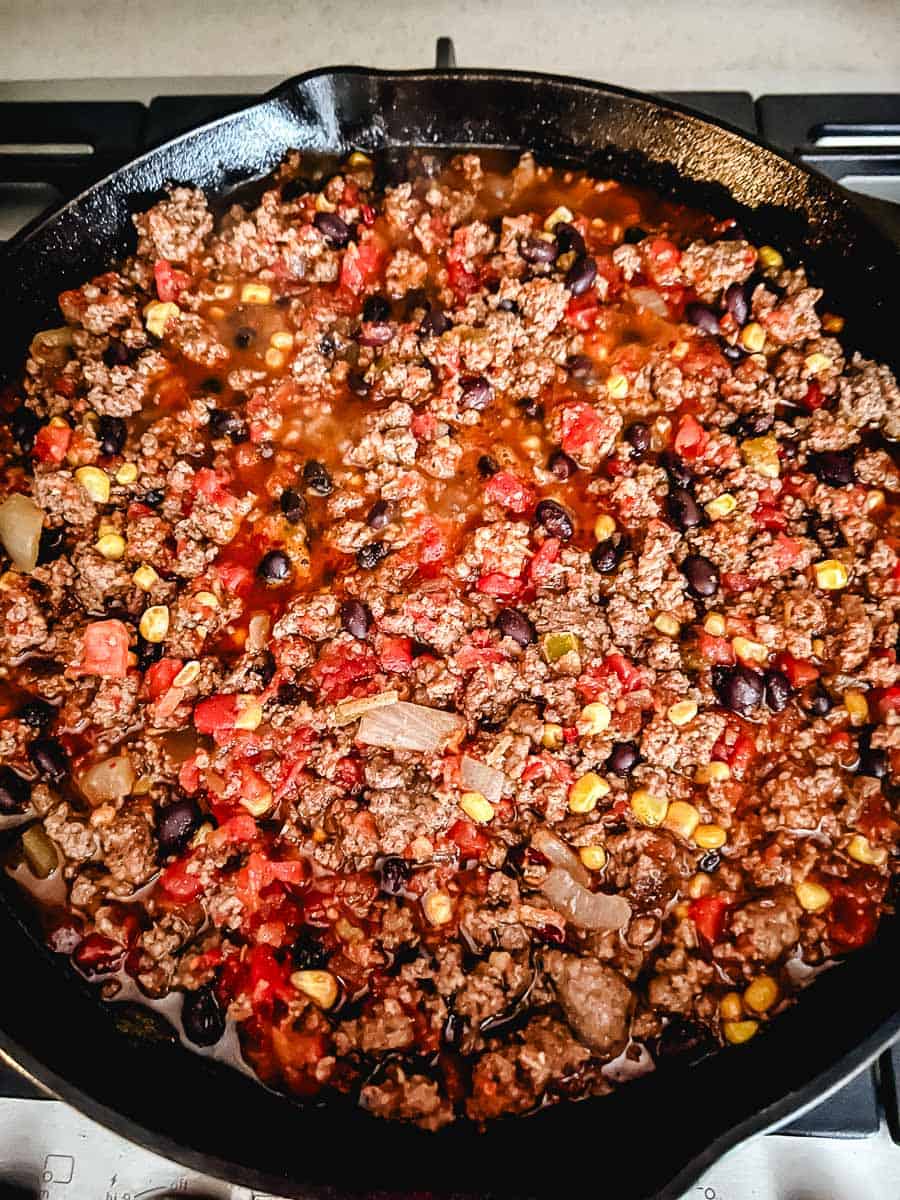 Ground beef taco skillet simmering with diced tomatoes, black beans, corn, and onion in a black cast iron pan on a gas stovetop