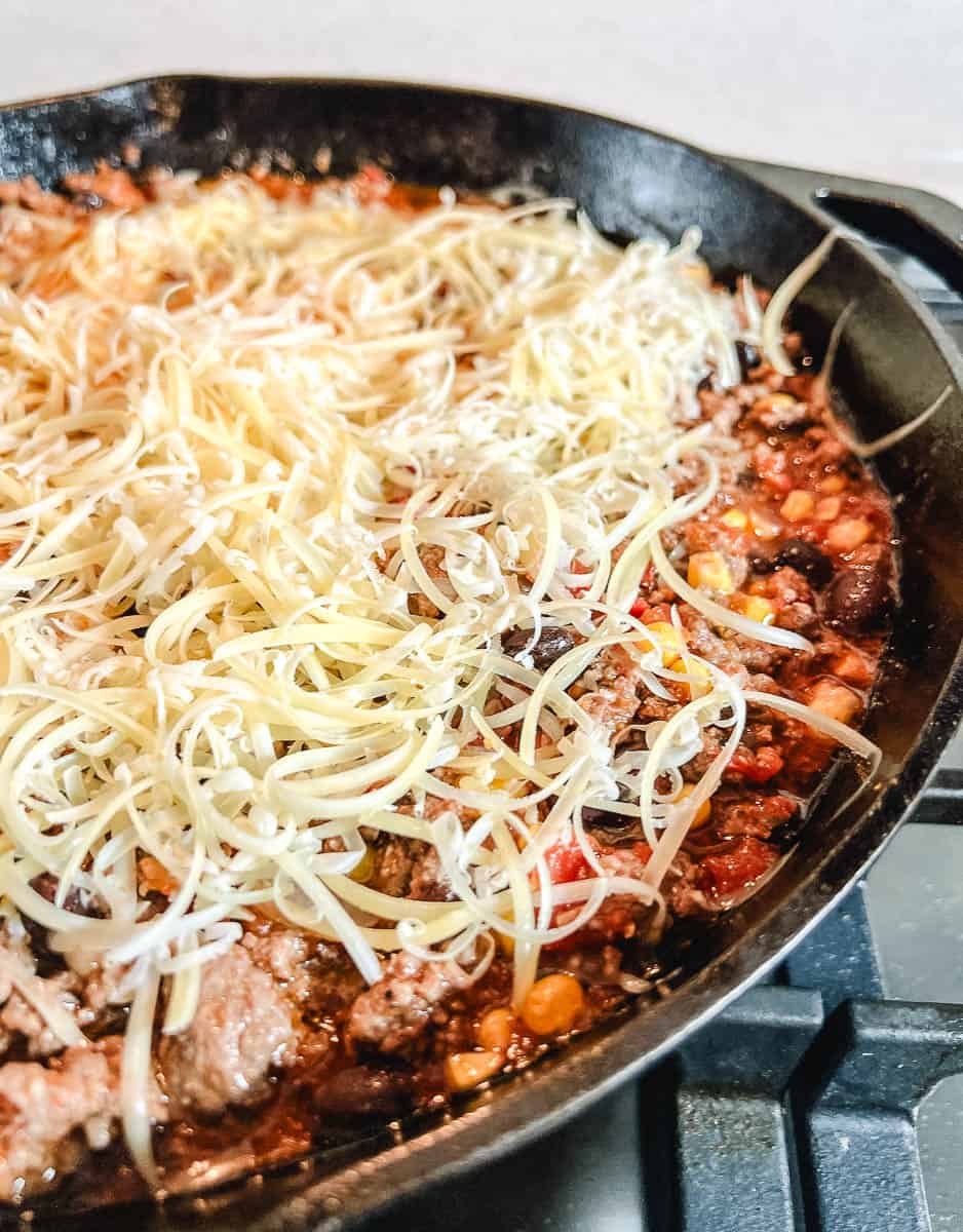 Shredded white cheese piled on top of a ground beef taco skillet dinner with black beans, corn, and tomatoes in a black cast iron skillet on a gas stovetop