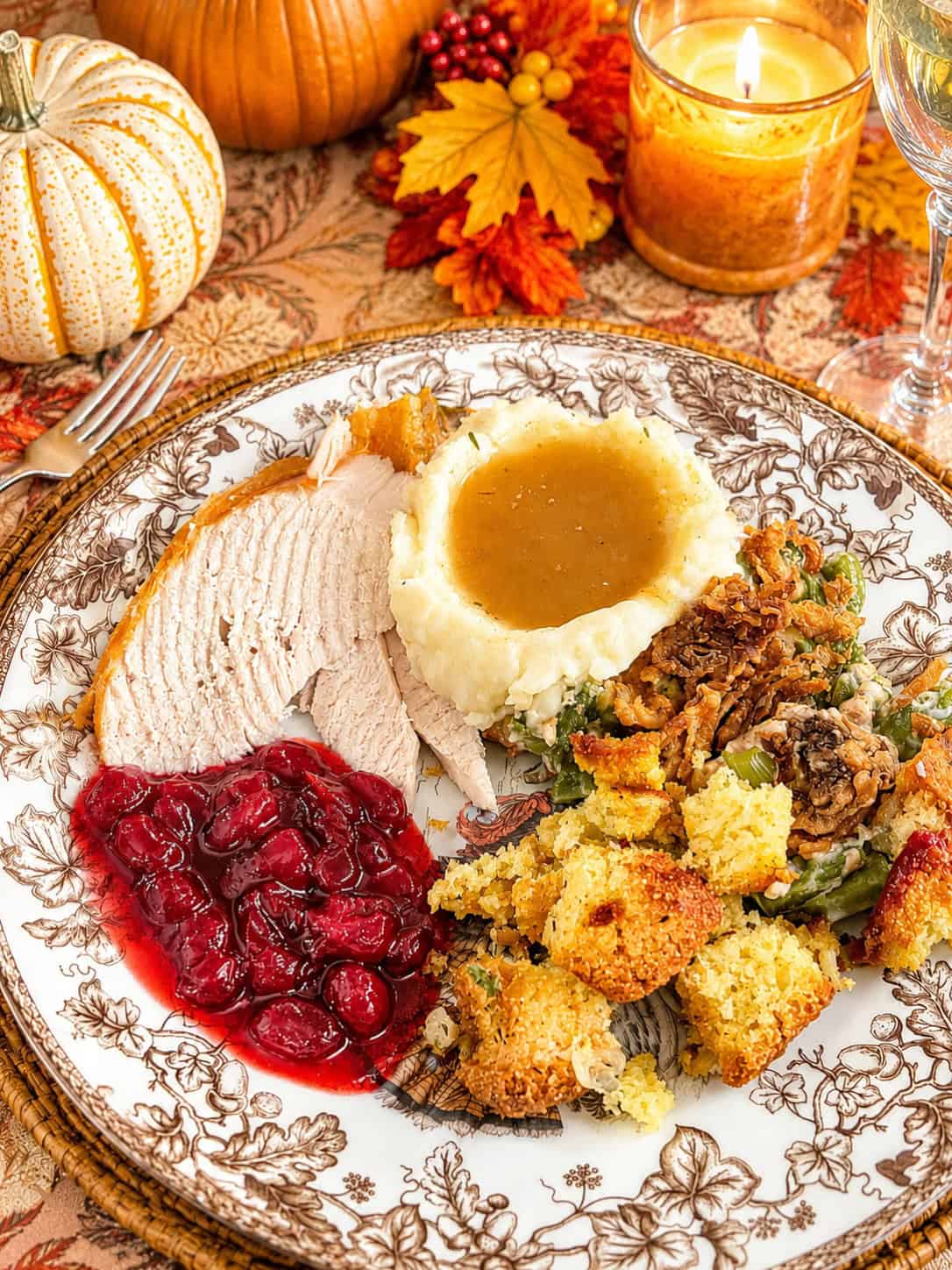 Homemade turkey gravy with drippings pooled over creamy mashed potatoes on a Thanksgiving plate with sliced turkey, cranberry sauce, and cornbread stuffing, set on a fall table with pumpkins and a glowing candle.