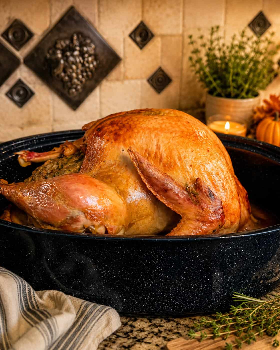 A golden roasted turkey resting in a black roasting pan full of drippings for homemade turkey gravy, with fresh thyme, a small pumpkin, and a lit candle on a rustic kitchen counter.
