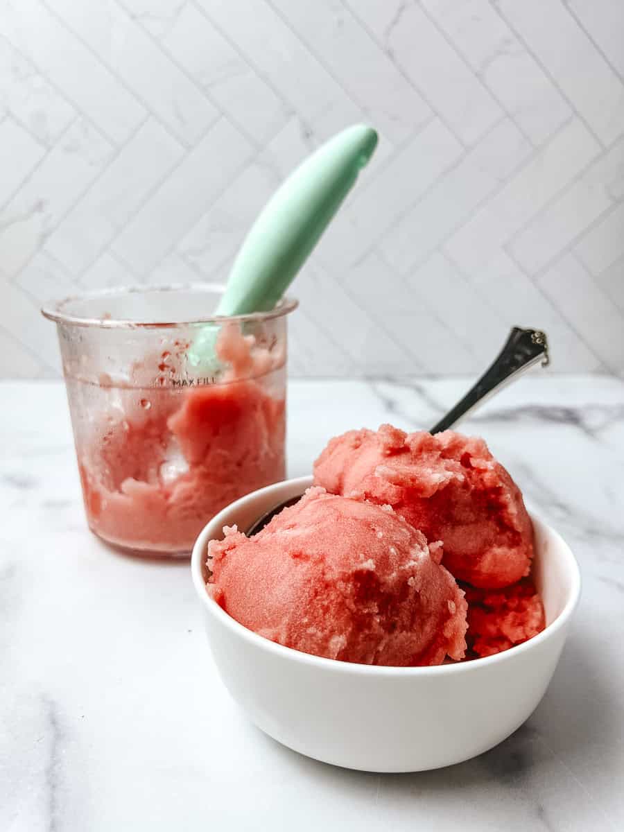 Scoops of watermelon sorbet in a white bowl next to a Ninja Creami pint with a mint green spoon on a marble counter with white tile behind.