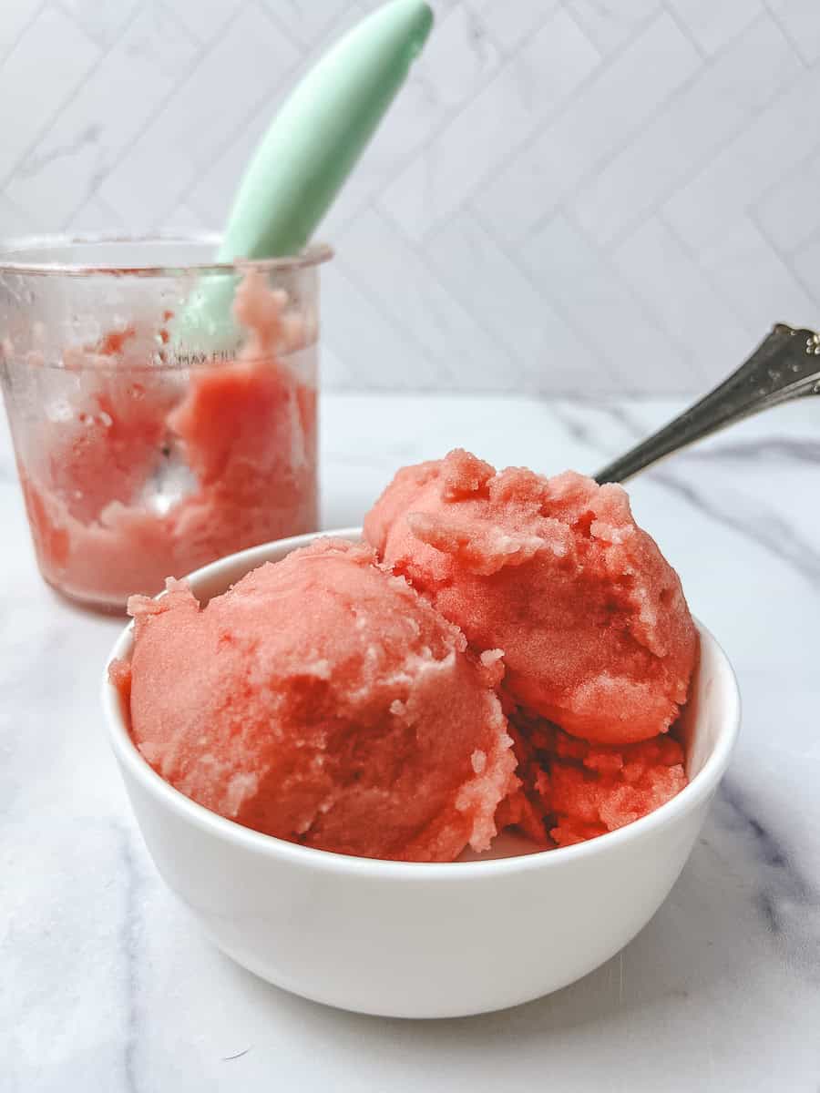 Ninja Creami watermelon sorbet scooped into a white bowl with the Creami pint and a mint green scoop behind it on a marble counter.