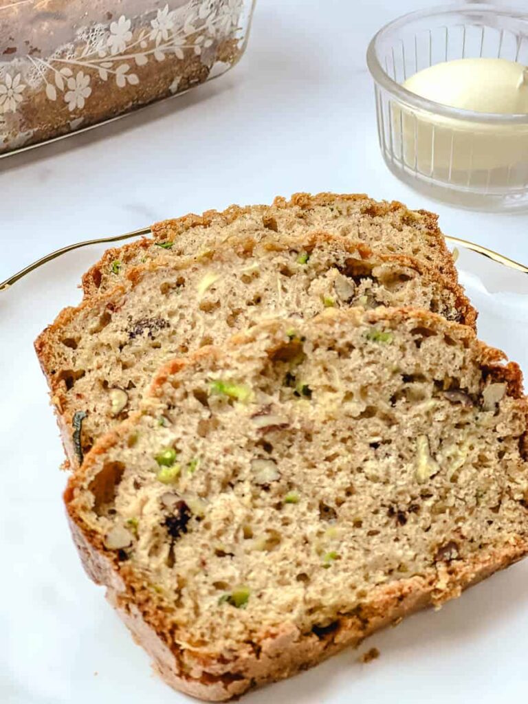 Three slices of low sugar zucchini bread on a white plate, showing a moist crumb with green zucchini flecks and chopped walnuts, with a small dish of butter and a floral loaf pan on a white marble counter.