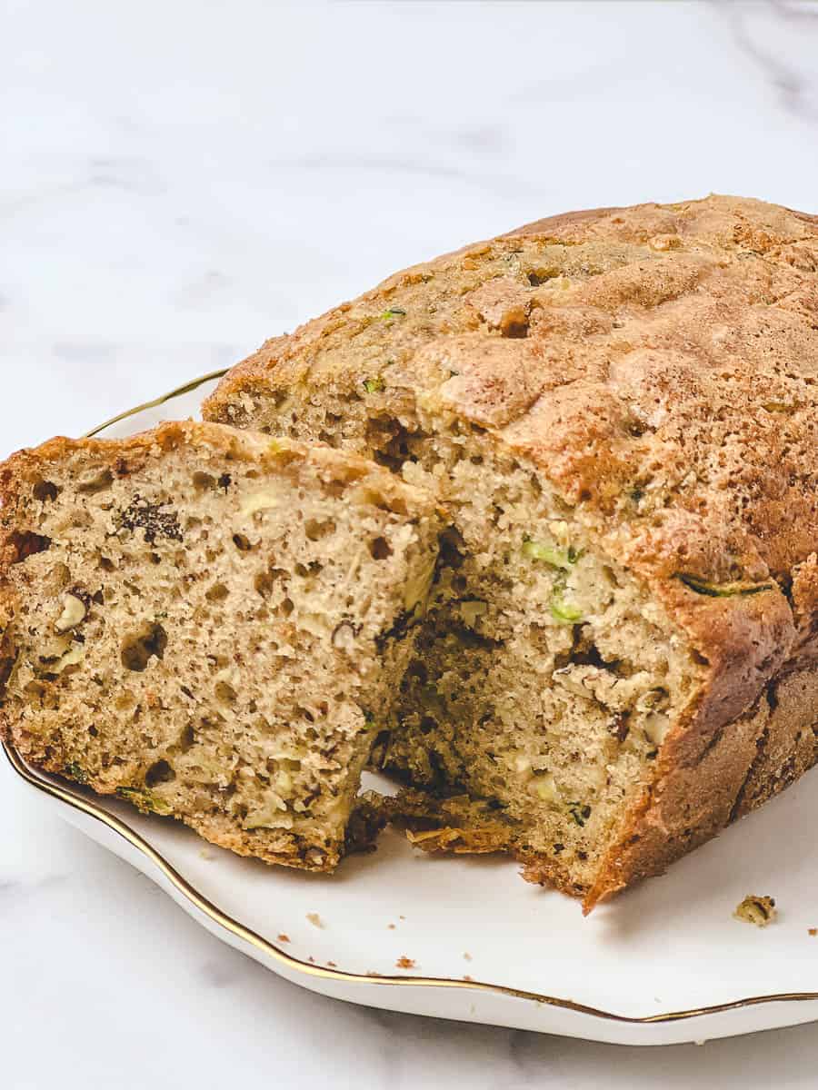 A sliced loaf of low sugar zucchini bread on a white gold-rimmed plate, showing the soft moist crumb with green zucchini flecks and walnuts inside, made with olive oil and no seed oils.