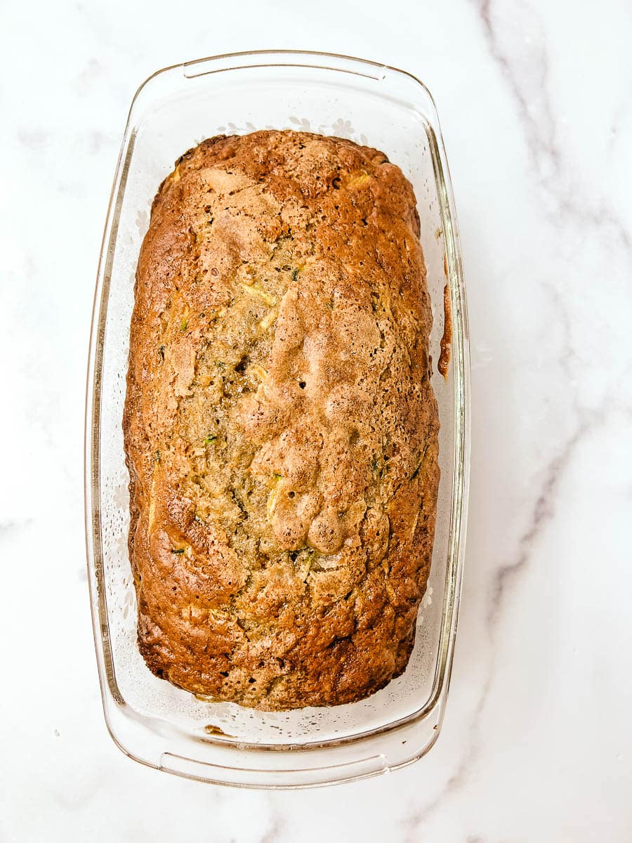Overhead view of a whole low sugar zucchini bread loaf baked in a clear glass pan on a white marble counter, with green zucchini flecks and a golden crackled top made with olive oil and no seed oils.