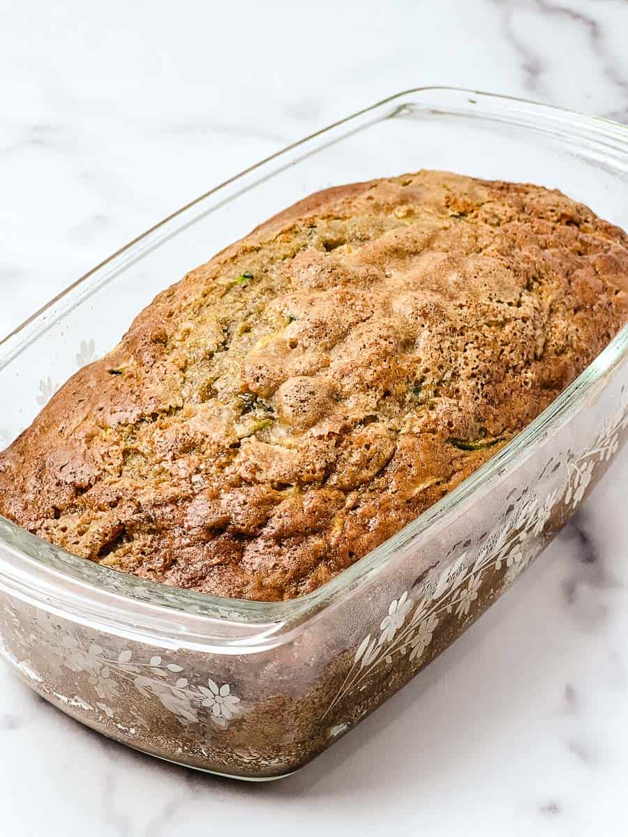 A whole loaf of low sugar zucchini bread with a golden domed top baked in a vintage floral glass loaf pan, cooling on a white marble counter.