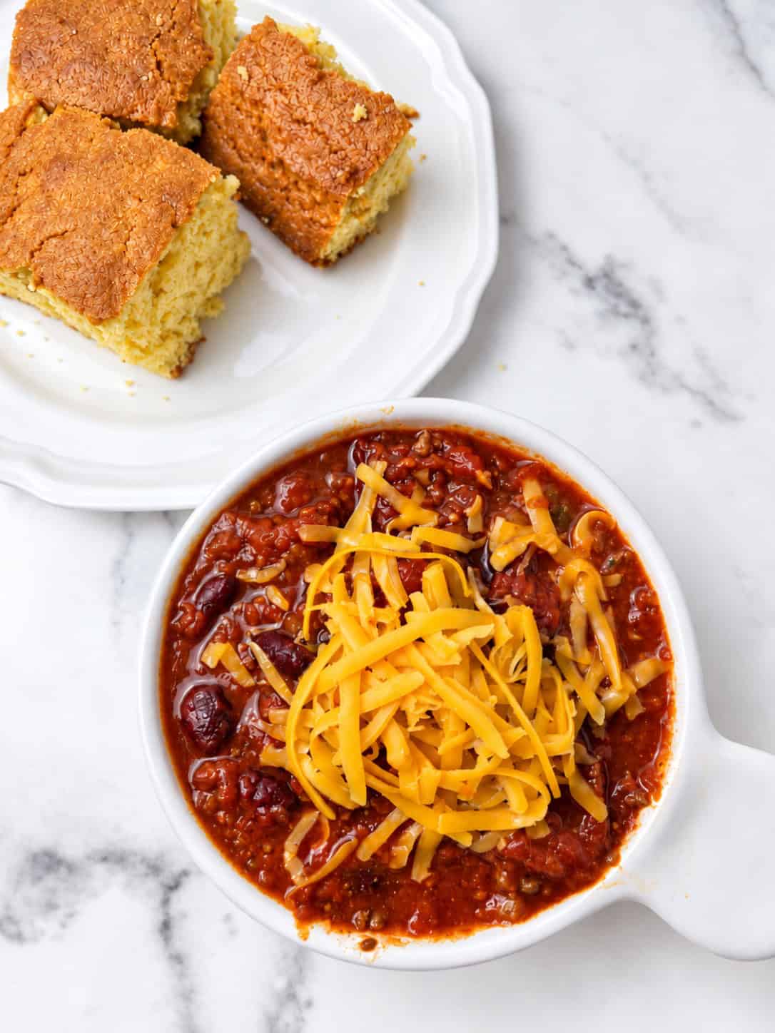 A white handled bowl of homemade beef chili with kidney beans, topped with shredded cheddar cheese and a silver spoon, with squares of golden cornbread on a white plate on a white marble counter.