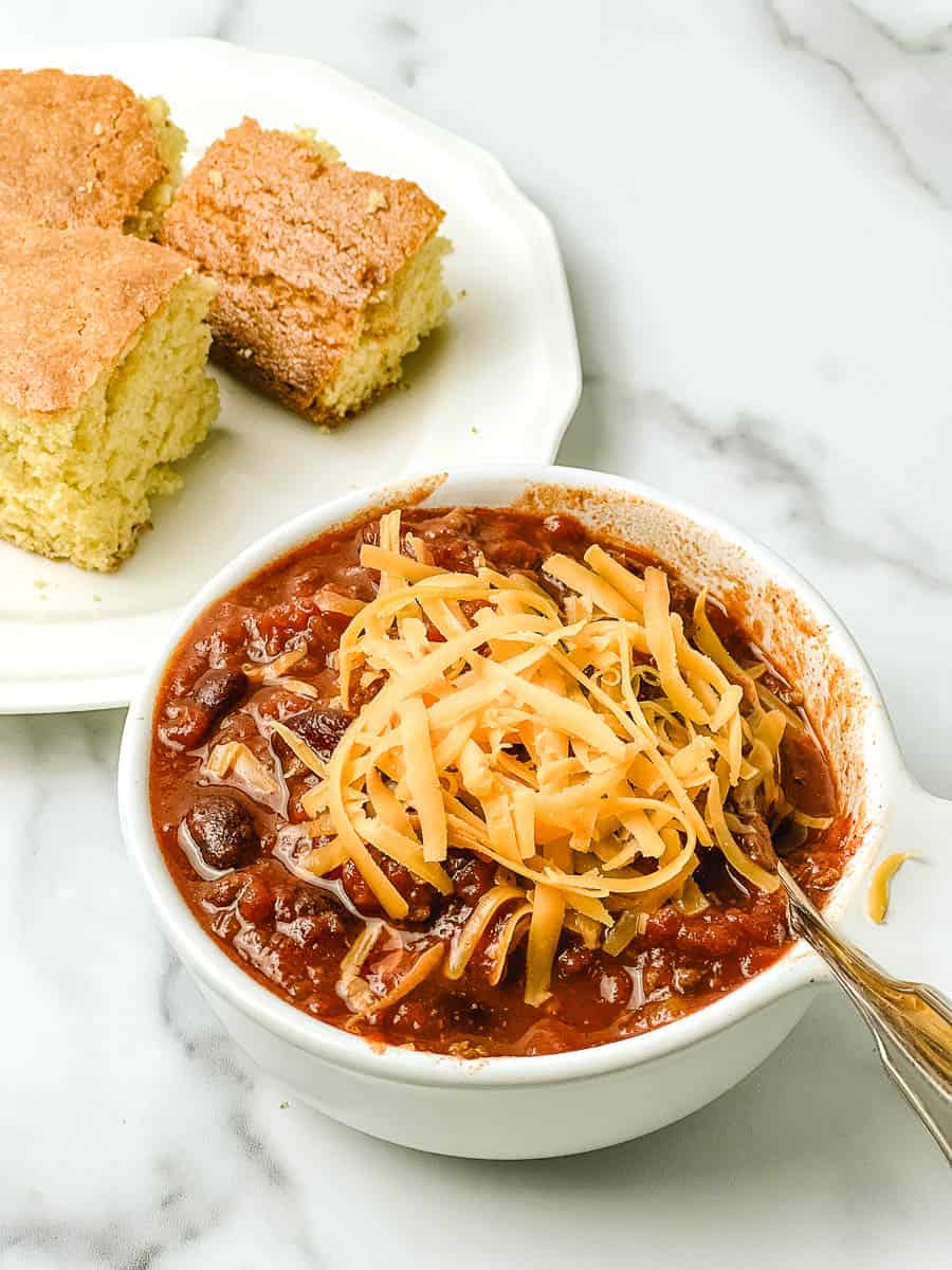 Easy crockpot chili with ground beef and kidney beans topped with shredded cheddar in a white bowl, with squares of homemade cornbread on a white plate on a marble counter.