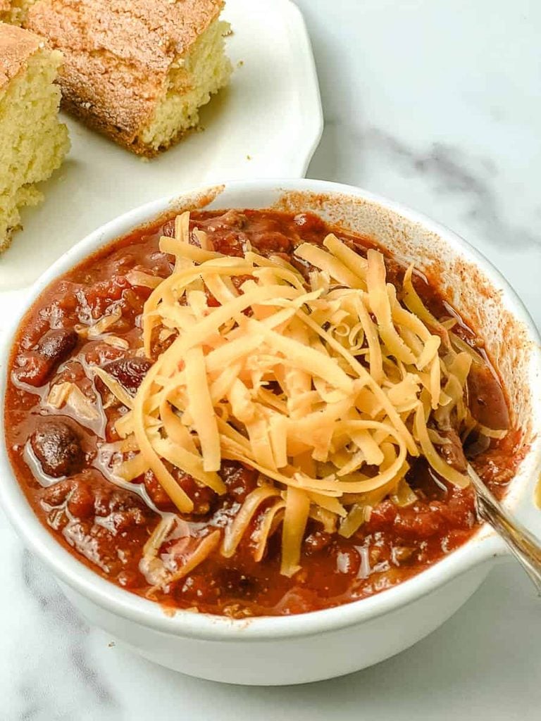 Close up of slow cooker chili with kidney beans and tomatoes topped with a pile of shredded cheddar in a white bowl, with homemade cornbread on the side.