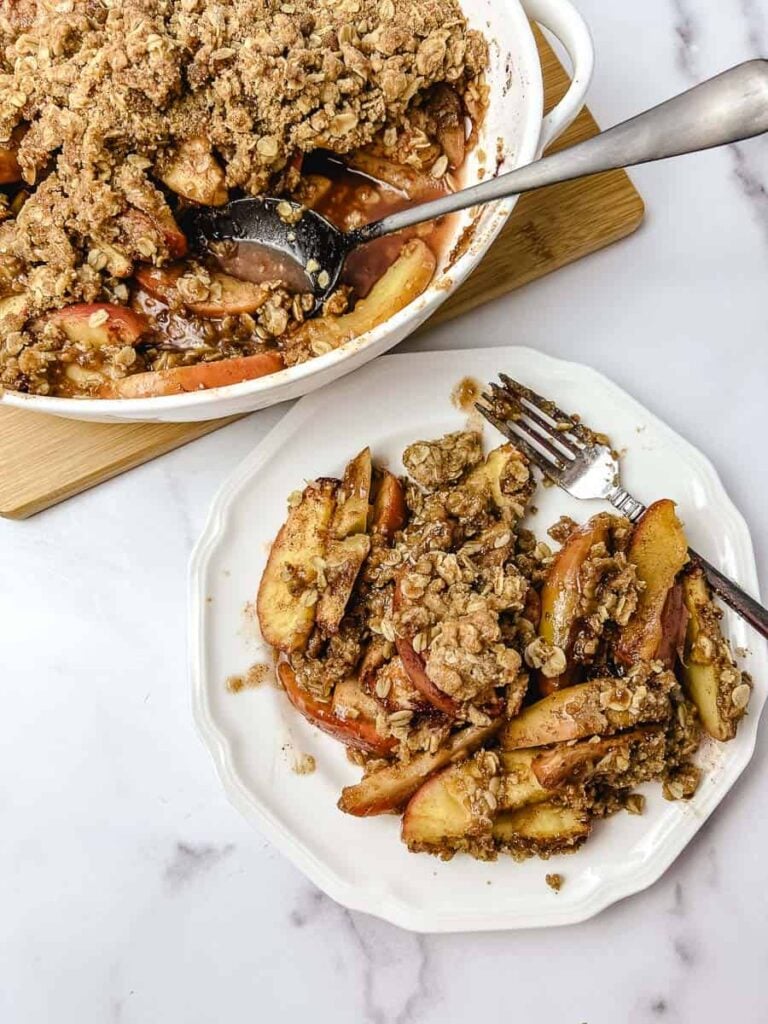 Homemade apple crisp with baked apple slices and oat crumble topping, served on a white plate beside the baking dish. top down view