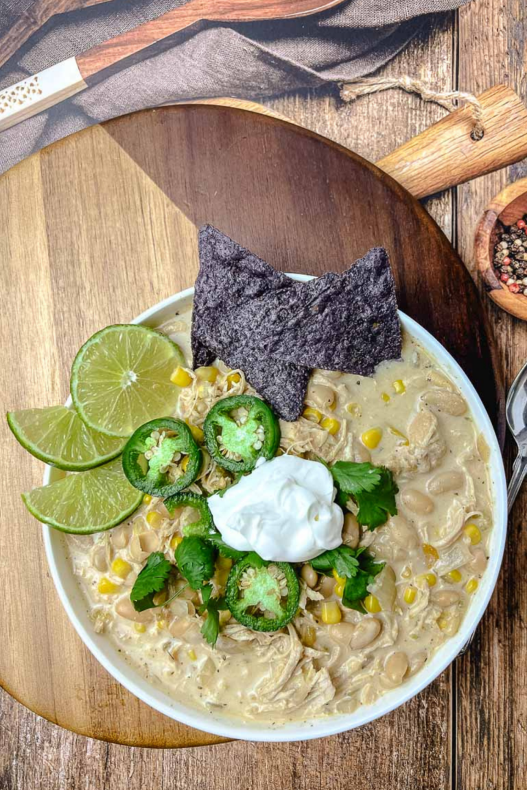 Overhead view of creamy slow cooker white chicken chili in a white bowl on a round wooden board, topped with sour cream, jalapeño slices, cilantro, lime wedges, and blue corn chips