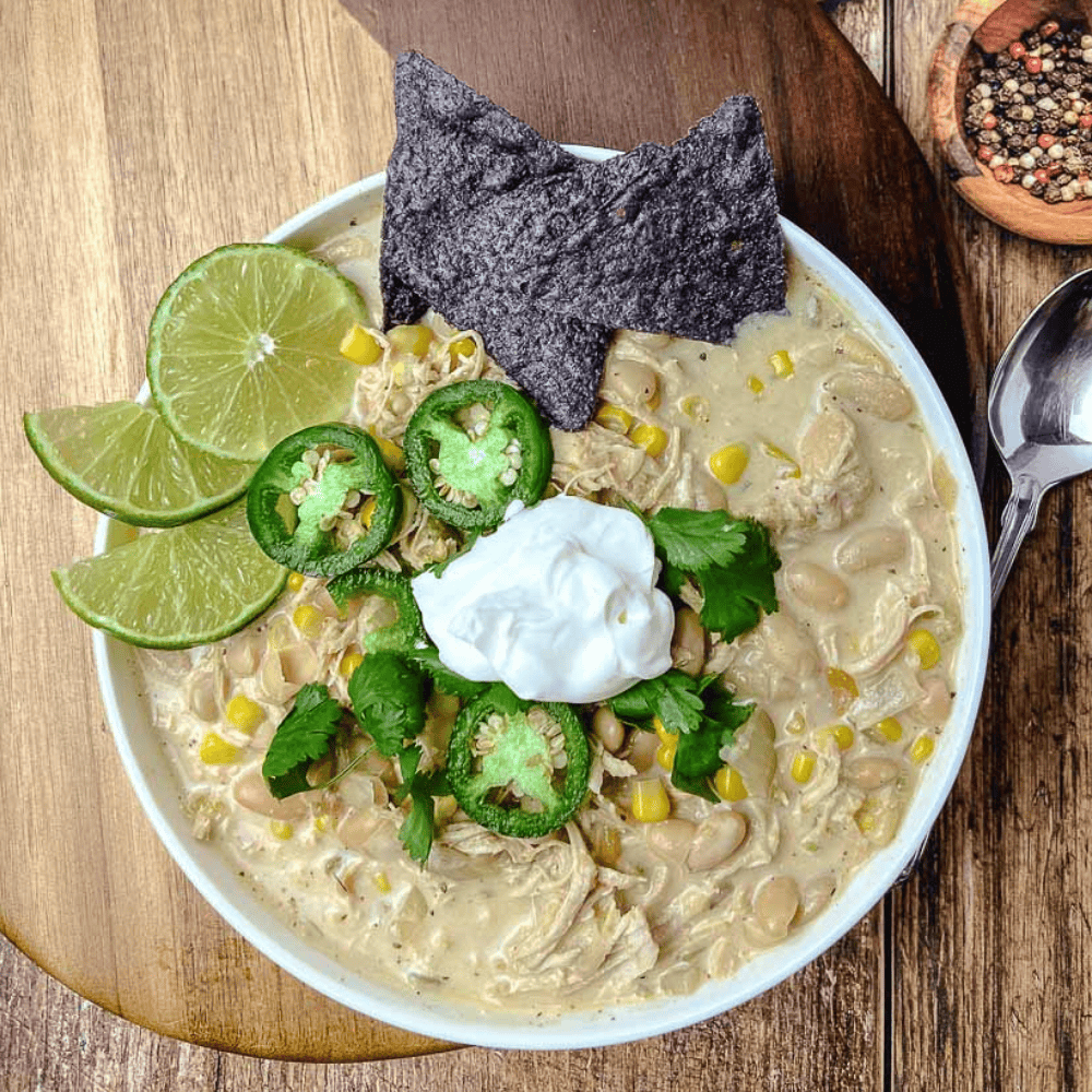 Square overhead photo of slow cooker white chicken chili in a white bowl, topped with sour cream, jalapeño slices, cilantro, lime wedges, and a blue corn chip, on a wooden board with a spoon and a small bowl of peppercorns