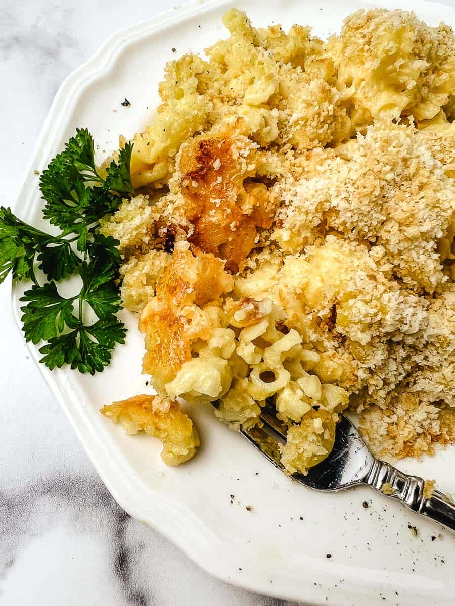 Close up of creamy baked mac and cheese on a white plate with a crispy panko top, a fork, and fresh parsley