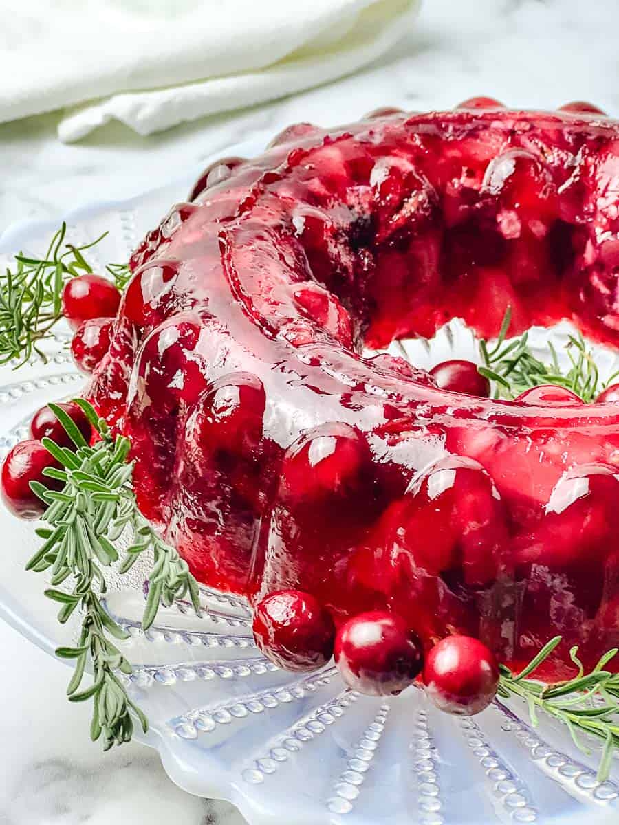 Close up of a glossy cranberry jello salad mold with whole cranberries and rosemary on a glass serving plate