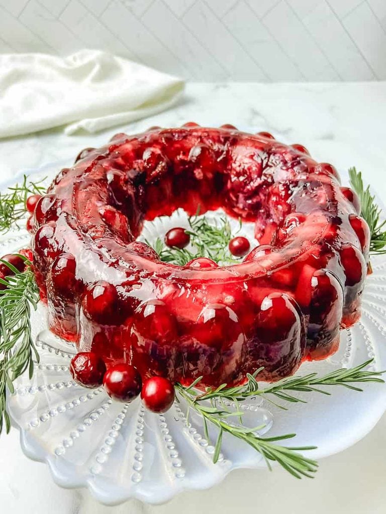 Cranberry jello salad mold turned out onto a clear glass plate with fresh cranberries and rosemary around the base, on white marble with a white napkin.